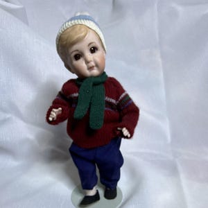 Puede incluir: Una muñeca de porcelana con cabello rubio que lleva un gorro de punto blanco y azul, una bufanda de punto verde oscuro, un suéter de manga larga color granate con un patrón de rayas horizontales multicolor, pantalones azul oscuro y zapatos negros.