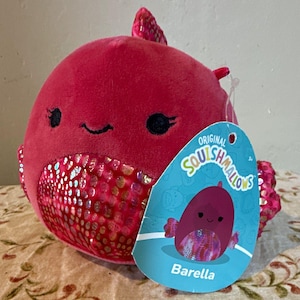 Pode incluir: Um brinquedo de pelúcia redondo em forma de peixe, com um corpo magenta vibrante e um padrão de escamas iridescentes e brilhantes na barriga e nas barbatanas. Possui olhos pretos bordados e um pequeno sorriso. Uma etiqueta azul diz Original Squishmallows Barella.