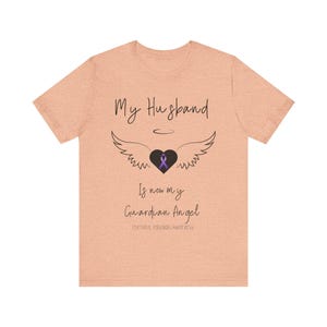 May include: A light peach t-shirt with the text "My Husband is now my Guardian Angel" and a purple ribbon inside a black heart with angel wings. The text "Fentanyl Poisoning Awareness" is also included.
