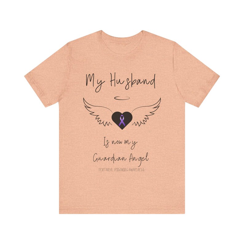 May include: A light peach t-shirt with the text "My Husband is now my Guardian Angel" and a purple ribbon inside a black heart with angel wings. The text "Fentanyl Poisoning Awareness" is also included.