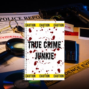 May include: A spiral-bound notebook with the text "TRUE CRIME JUNKIE" in black, splattered with red ink. Yellow caution tape borders the top and bottom. The background includes a police report, a handgun, keys, and a magnifying glass.