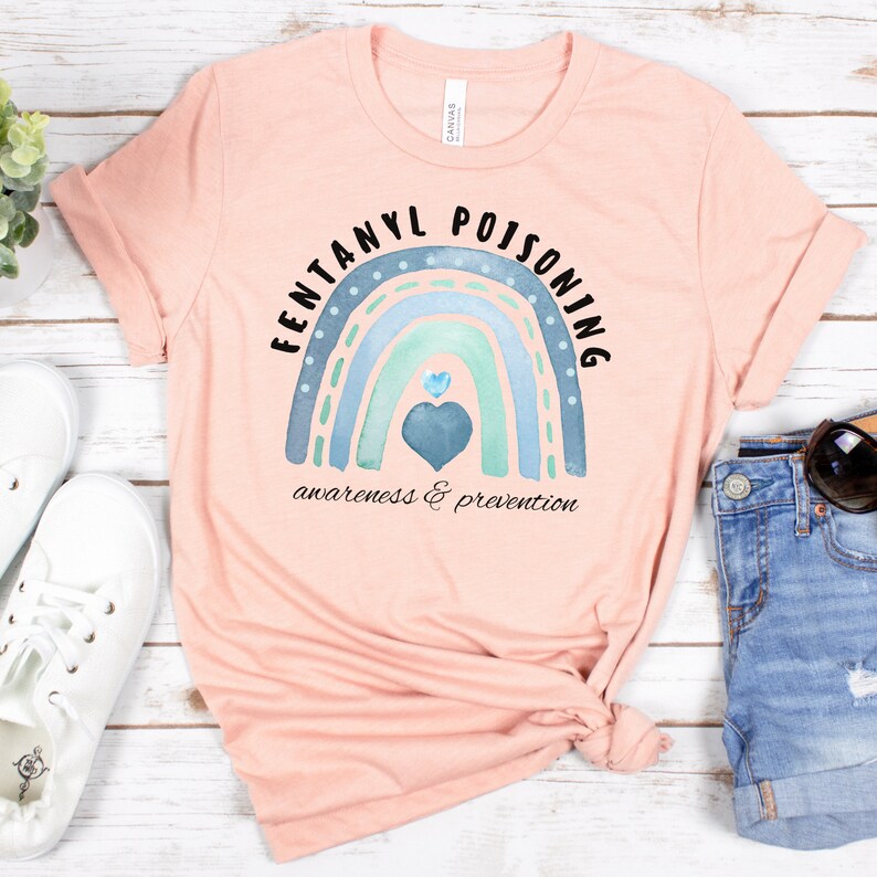 Puede incluir: Una camiseta rosa con un dise&ntilde;o de arco&iacute;ris azul y verde y el texto "Fentanyl Poisoning awareness & prevention".