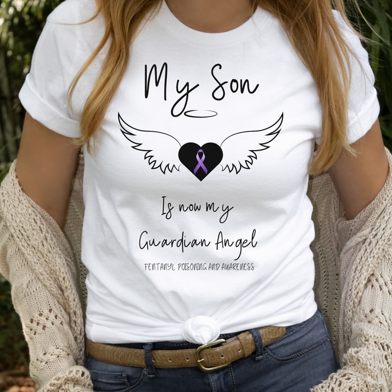 May include: White t-shirt with the text "My Son Is now my Guardian Angel" and "Fentanyl Poisoning and Awareness". The design includes a black heart with a purple ribbon, angel wings, and a halo.