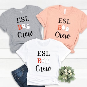 Könnte beinhalten: Drei T-Shirts mit dem Text "ESL Boo Crew" in schwarzer Schrift mit einem roten "B" und zwei weißen Geisterillustrationen. Die Shirts sind in Grau, Pfirsich und Weiß.