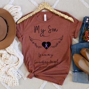 May include: A rust-colored t-shirt with the words "My Son" above a heart with wings and a purple ribbon. Below the heart, the text reads "Is now my Guardian Angel" and "Fentanyl Poisoning and Awareness."