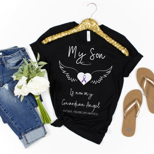 May include: Black t-shirt with the text "My Son Is now my Guardian Angel" and a graphic of wings, a heart, and a purple ribbon. The shirt is displayed on a gold hanger, with jeans, flowers, and sandals in the background.
