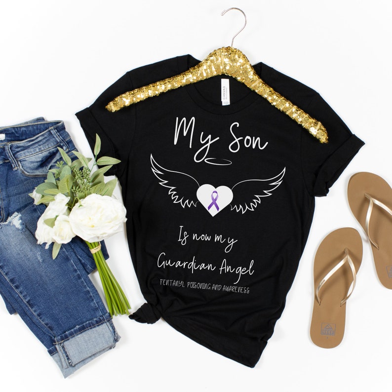 May include: Black t-shirt with the text "My Son Is now my Guardian Angel" and a graphic of wings, a heart, and a purple ribbon. The shirt is displayed on a gold hanger, with jeans, flowers, and sandals in the background.
