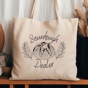 May include: A white canvas tote bag with black text that reads "Sourdough Dealer" and a black and white illustration of a sourdough loaf of bread with leaves on either side.
