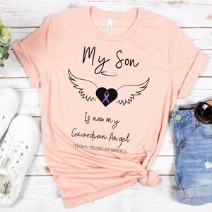 May include: A light pink t-shirt with the text "My Son Is now my Guardian Angel" in black script. A black heart with a purple ribbon and angel wings is centered above the text. The bottom text reads "Fentanyl Poisoning and Awareness".