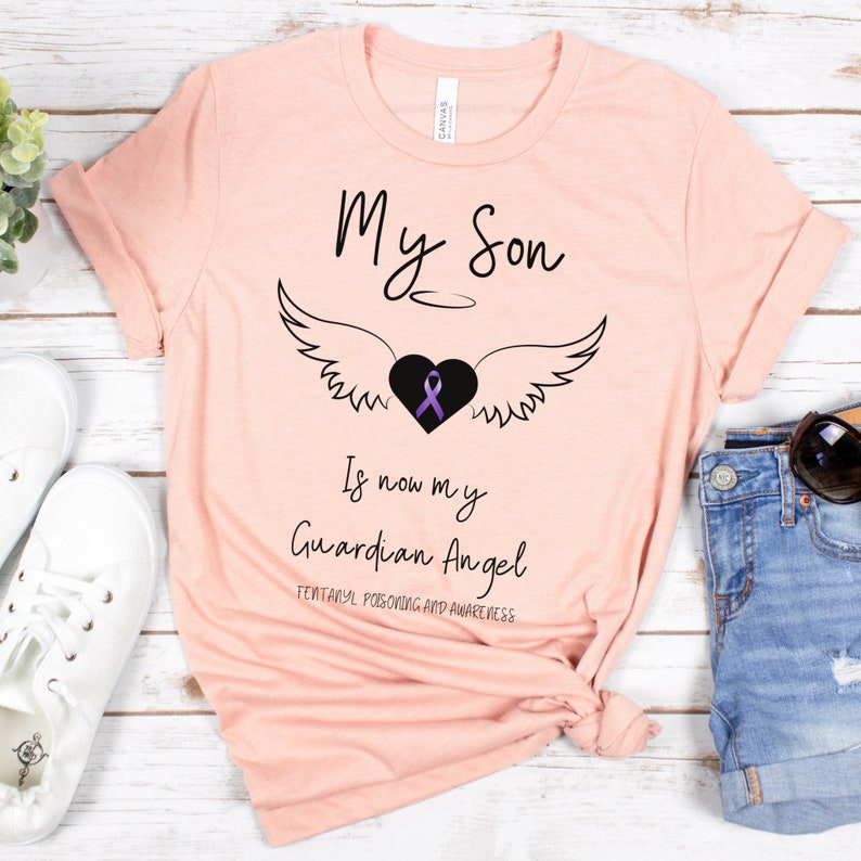 May include: A light pink t-shirt with the text "My Son Is now my Guardian Angel" in black script. A black heart with a purple ribbon and angel wings is centered above the text. The bottom text reads "Fentanyl Poisoning and Awareness".