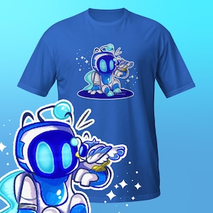 May include: Royal blue t-shirt featuring a cartoon robot character in shades of blue and white, holding a small bird. The design includes stars and a light blue background. The robot has a rounded head and a friendly expression.