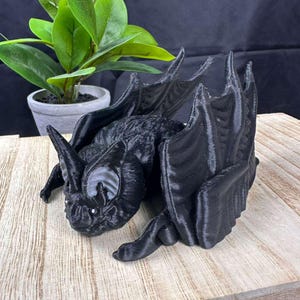 May include: A black, 3D-printed bat figurine with large wings, pointed ears, and detailed facial features. The bat is posed on a wooden surface, with a potted plant in the background. The figurine has a textured body and is a decorative item.