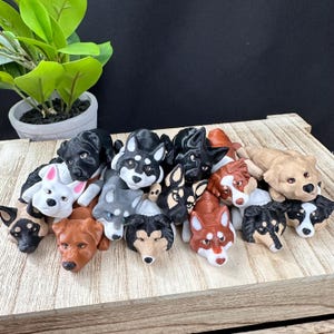 Pode incluir: Uma coleção de 16 pequenas estatuetas de cães impressas em 3D, de diferentes raças e cores. As estatuetas estão dispostas em uma pilha sobre uma superfície de madeira.