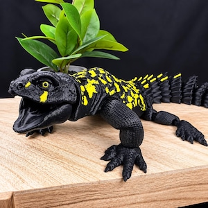 May include: An articulated lizard figurine featuring a black body with bright yellow spots and a textured, scaly surface. The creature has an open mouth, visible yellow eyes, and a segmented, spiky tail that extends behind its four clawed limbs.