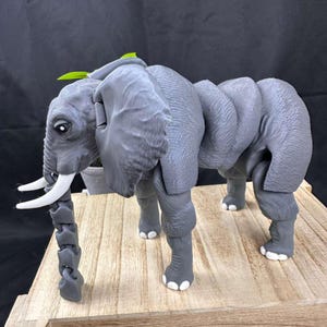 May include: A gray articulated elephant figurine with white tusks and a segmented trunk. The elephant has large ears and a green leaf on its head. The figurine is standing on a wooden surface.