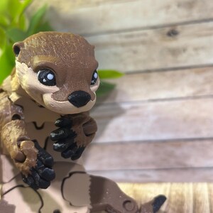 May include: A brown and tan articulated toy otter with black eyes and a black nose. The otter is sitting with its paws tucked in front of it.