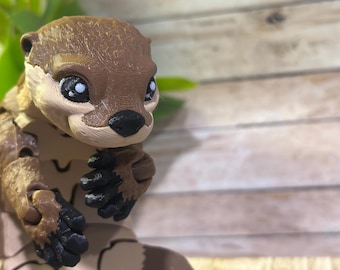 May include: A brown and tan articulated toy otter with black eyes and a black nose. The otter is sitting with its paws tucked in front of it.