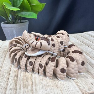 May include: A tan and brown articulated snake toy with a detailed design. The snake has brown spots and a detailed head with an orange and brown eye. The toy is coiled on a wooden surface.
