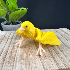 Puede incluir: Un pájaro de juguete de plástico amarillo con una superficie texturizada detallada. El pájaro tiene un pequeño pico y pequeñas patas. Las alas están extendidas y el pájaro está sentado sobre una superficie de madera.