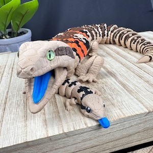 May include: A detailed 3D-printed gecko sculpture with a large adult and a smaller juvenile. The adult gecko features a tan body with brown and orange stripes, green eyes, and a blue tongue. The juvenile has black markings and a blue tongue.