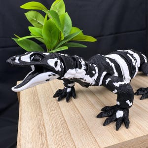 May include: An articulated lizard figurine featuring a high-contrast black and white mottled pattern. The lizard is depicted with its mouth open, showing a white interior, and has a segmented body structure with detailed claws on each foot.