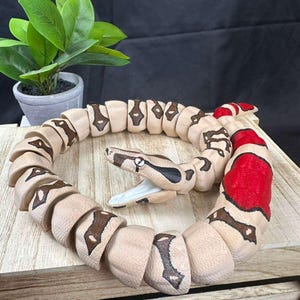 May include: A wooden snake toy with a tan body and brown markings. The snake's head is detailed with black and brown patterns, and its tail features red accents. The toy is coiled on a wooden surface.