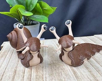 May include: Two brown and tan snail figurines with large, expressive eyes. The snails have detailed shells and bodies, and are posed on a wooden surface. A potted plant is in the background.