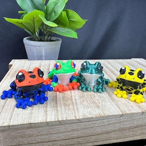 May include: Four colorful frog figurines in a row. The frogs are in various colors: orange and blue, green and red, teal, and yellow and black. The frogs are sitting on a wooden surface.
