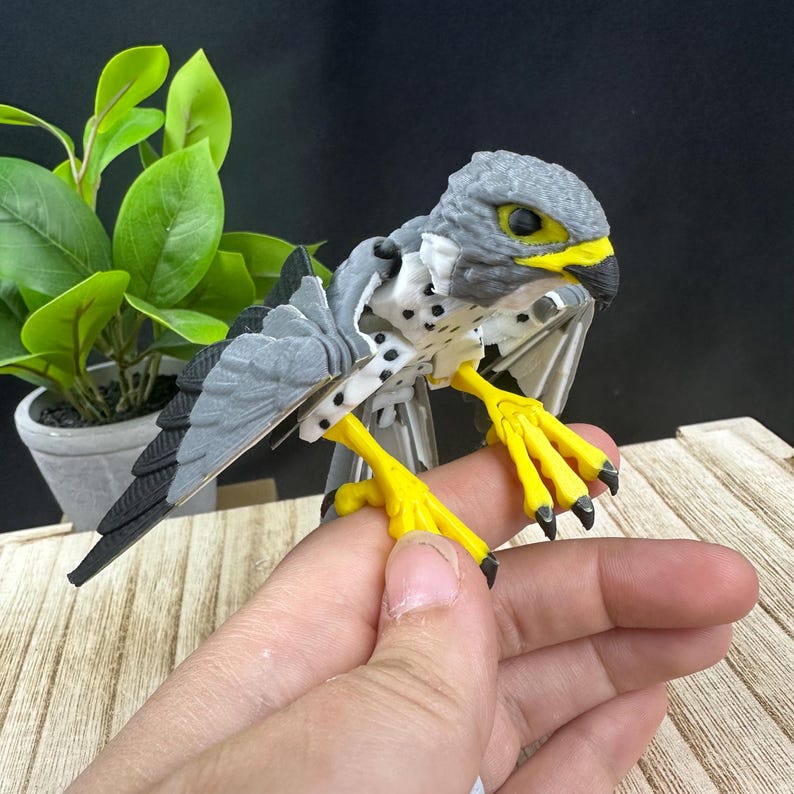 May include: A gray and white 3D printed model of a falcon with yellow legs and black talons. The model is posed with its wings spread and is perched on a finger.