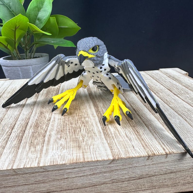 May include: A gray and white 3D printed model of a falcon with yellow talons and black wing tips. The falcon is posed with its wings spread and its head turned to the side.