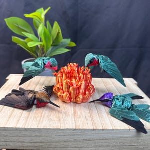 May include: Four colorful hummingbird figurines with outstretched wings surround a vibrant orange and gold decorative object. The hummingbirds have green, brown, and purple accents. The scene is set on a wooden surface with a plant in the background.