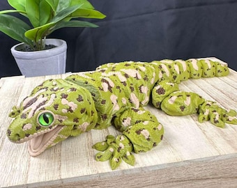 May include: A detailed, green and tan gecko figurine with an open mouth and a bright green eye. The reptile has a spotted pattern and is posed on a wooden surface. A small potted plant is in the background.