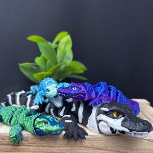 May include: Four toy alligators in different colors and patterns. A green alligator, a blue and purple alligator, a black and white alligator, and a blue alligator.
