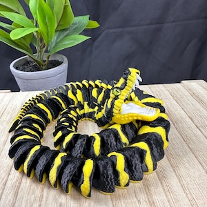 May include: A 3D-printed articulated snake toy in black and yellow. The snake is coiled, with its head raised, revealing white teeth and a yellow mouth. The snake's body has a repeating pattern of black and yellow stripes. A green-eyed snake is on a wooden surface.