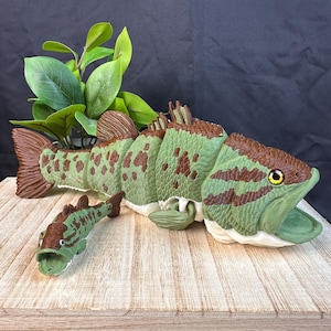 May include: Two articulated fish figures featuring a green and brown mottled scale pattern. The larger fish has a wide-open mouth and yellow eyes, while the smaller fish is a miniature version of the same design with a similar color scheme.