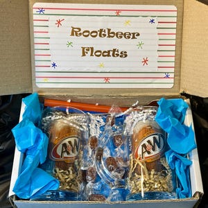 May include: A gift box containing two A&W root beer glasses, candies, and straws. The glasses are filled with shredded paper and wrapped in blue tissue paper. The box lid has a sign that reads "Rootbeer Floats".