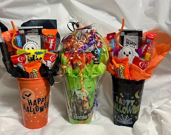 May include: Three Halloween-themed plastic cups filled with assorted candy and snacks. The orange cup reads Happy Halloween, the green cup features a haunted house and reads My Hauntings, and the black cup reads Happy Halloween with a spiderweb.
