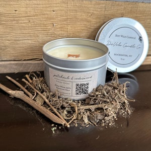 Op de afbeelding: Een zilveren kaarsblik met een witte sojawas kaars erin. De kaars is gelabeld met "patchouli & cederhout" en "Gemaakt met 100% etherische oliën". Het blik heeft een houten deksel en een QR-code op het etiket. De kaars staat op een bed van gedroogde kruiden.