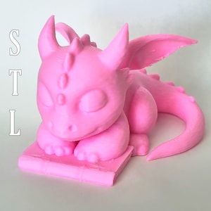 May include: A pink 3D printed dragon figurine sleeping on a pink book. The dragon has closed eyes and a curled tail. The book has a textured surface.