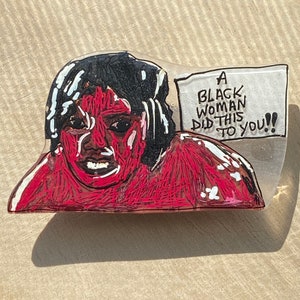 May include: A hand-painted illustration of a person with a red shirt and black hair. The person is looking at the viewer with a surprised expression. A speech bubble above the person's head reads "A BLACK WOMAN DID THIS TO YOU!!"