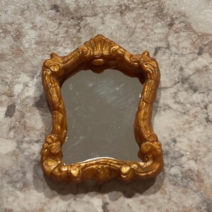 May include: A small, ornate gold-colored mirror with a decorative frame. The mirror has a rectangular shape and is surrounded by intricate details.