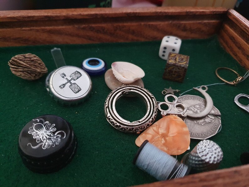 May include: A wooden box filled with various trinkets, including a silver ring, a seashell, a bottle cap with a graphic of a shovel, a small silver charm with three circles, a silver crescent moon charm, a silver star charm, a small silver thimble, a small silver coin with the word "Australia" on it, a small blue glass eye, a small brown nut, a small white die, a small brown metal object with a design on it, and a small white plastic object.