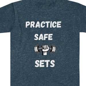 Op de afbeelding: Een donkerblauw T-shirt met de tekst "PRACTICE SAFE SETS" in wit. Een zwart-witte illustratie van een hand die een halter optilt, bevindt zich tussen de woorden "SAFE" en "SETS".