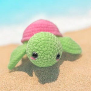 May include: A green and pink crocheted turtle toy with black eyes and pink cheeks. The turtle is lying on its belly on a sandy surface.