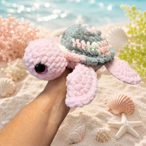 May include: A plush turtle toy crafted from soft, chunky yarn in shades of light pink and dark gray. The turtle features a rounded head with a large, glossy black eye and four flippers. The shell displays a textured pattern of gray and pink.
