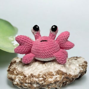May include: A small, handmade crochet crab toy crafted from soft pink yarn. The creature features two prominent, stalk-like eyes with black pupils, a small embroidered mouth, and multiple textured pincers extending from its rounded body.