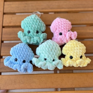 May include: Five handmade crochet octopus keychains in various pastel colors: light blue, mint green, pink, yellow, and a darker mint green. Each octopus has black button eyes and a silver keychain ring. The octopuses are displayed on a wooden surface.