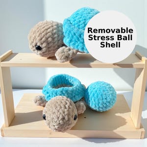 May include: Crocheted turtle stress ball with a removable shell. The turtle has a tan head and limbs, a light blue shell, and black eyes. The text "Removable Stress Ball Shell" is displayed on a white circle.