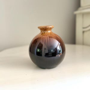 May include: A small, round ceramic vase with a glossy finish. The vase transitions from a dark brown base to a lighter brown at the top, with a textured rim. The vase is approximately 5 inches tall.