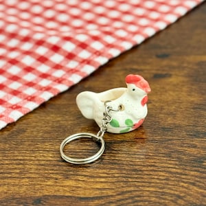 May include: A ceramic chicken keychain with a red comb, green and pink floral accents, and a silver keyring. The keychain is set against a wooden surface with a red and white checkered cloth.